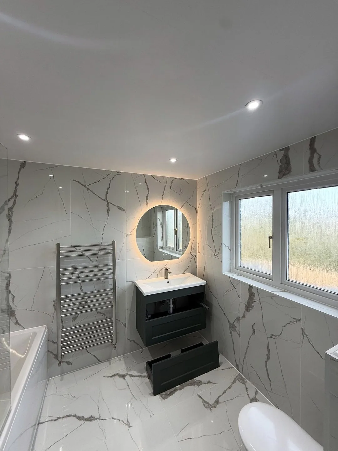 Marble bathroom with illuminated round mirror, vanity unit and heated towel rail