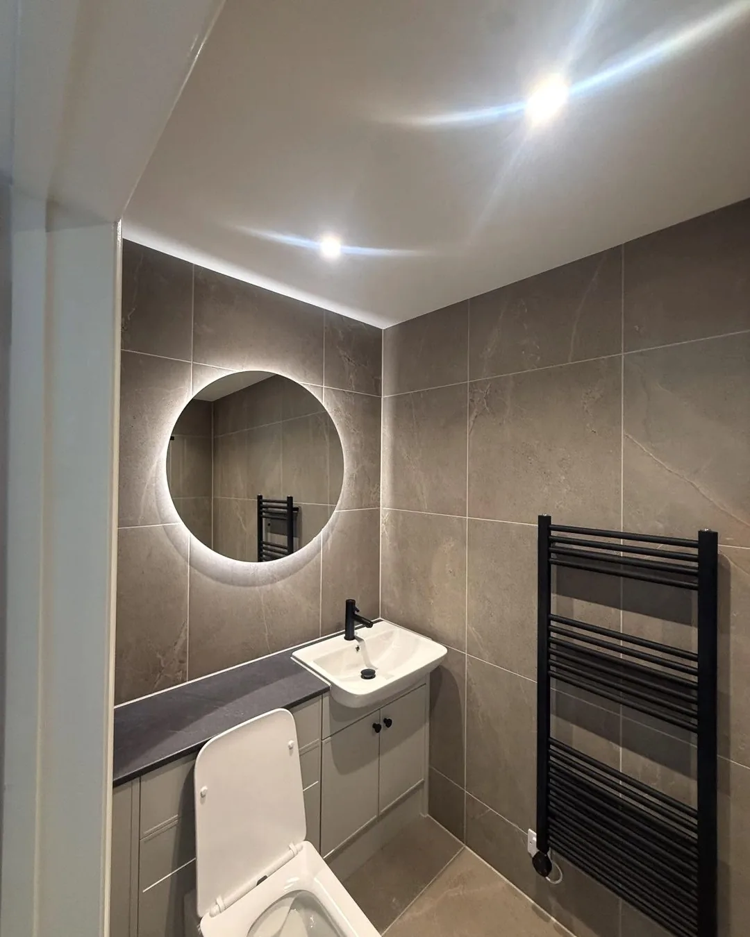 Tiled bathroom with illuminated round mirror, vanity basin and heated towel rail